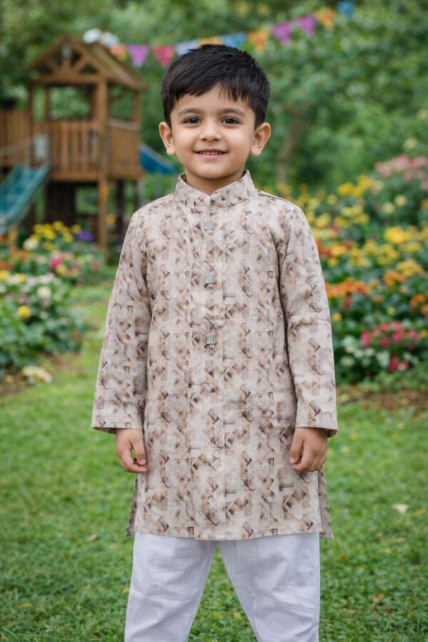 Boy wearing beige abstract printed kurta with white pajama, smiling outdoors in a garden setting.