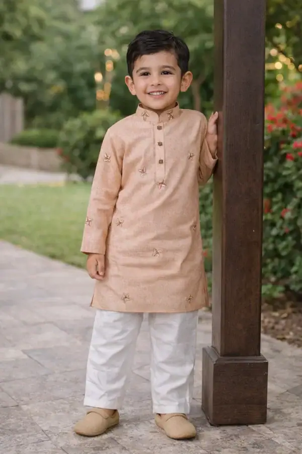 Cute boy wearing peach embroidered kurta with white pajama, smiling outdoors.